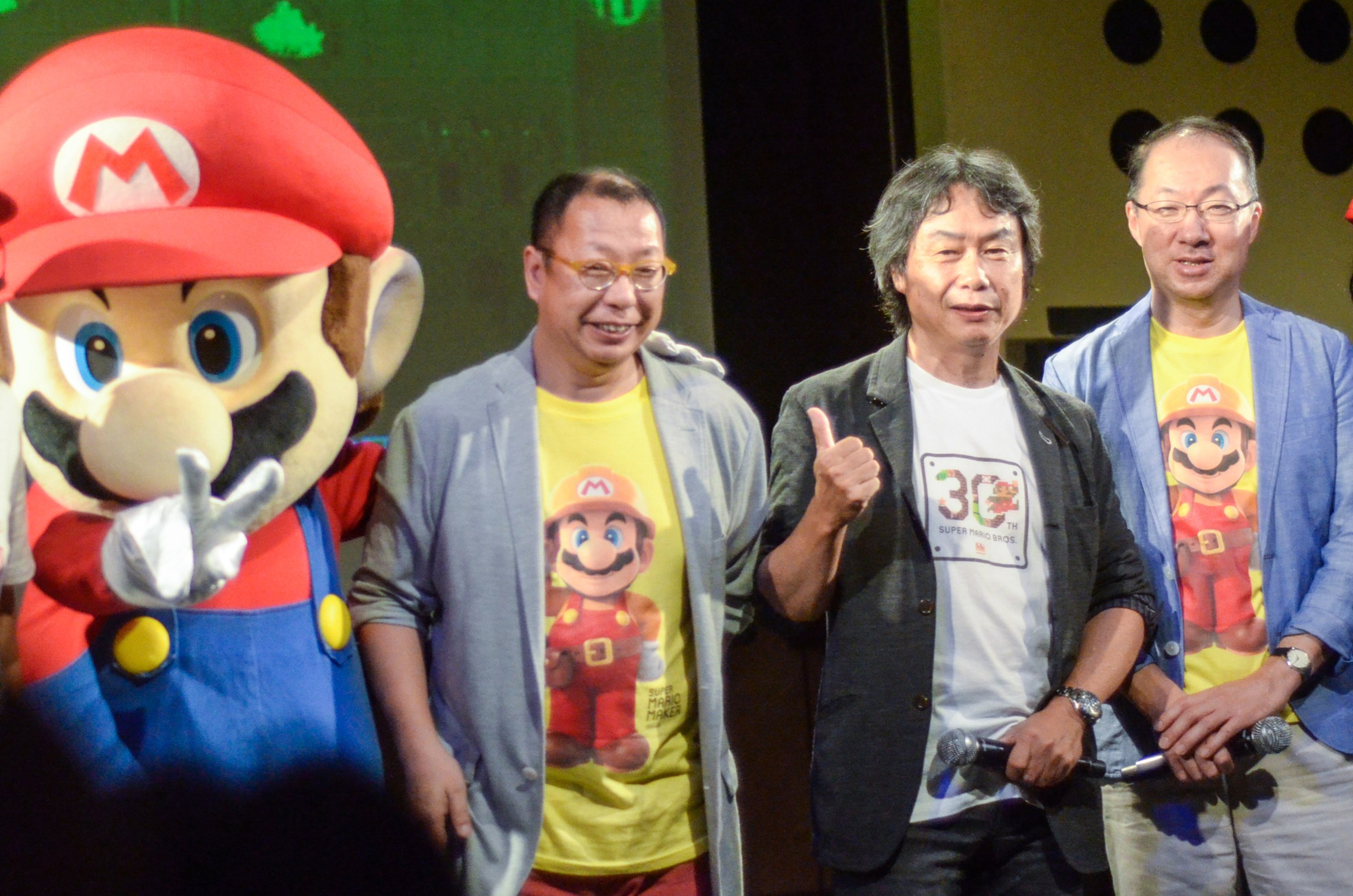 Three legendary Nintendo creators stand together at the Super Mario 30th Anniversary Festival. From left to right: A person in a life-sized Mario costume with red cap and blue overalls; Takashi Tezuka wearing glasses and a gray jacket over a yellow Mario t-shirt; Shigeru Miyamoto in the center wearing a black jacket over a white 30th anniversary t-shirt, giving a thumbs up; and Koji Kondo on the right in a blue jacket over a yellow Mario t-shirt. All three human creators are smiling warmly, embodying 30 years of bringing joy to millions. The Mario character costume towers over them, a reminder of how their creation has grown larger than life.