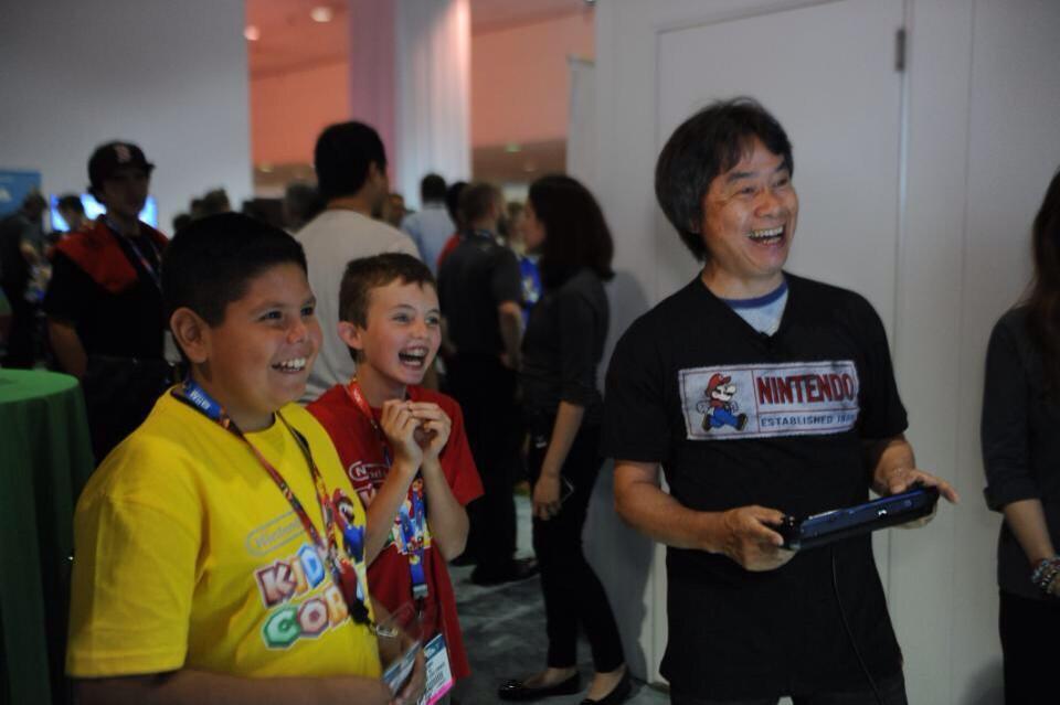 The architect of joy becomes the joyful observer Shigeru Miyamoto at what appears to be a gaming event, wearing his signature black Nintendo t-shirt with Mario graphics. He's surrounded by young fans - two boys visible in the frame, one in a yellow shirt and one in red, both grinning with excitement. Miyamoto holds a blue game controller and his face radiates genuine warmth and joy. His expression mirrors the grandfather from his keynote - not playing the game, but delighting in the happiness around him. This is the Wife-O-Meter at its ultimate reading: when the designer himself becomes the joyful observer of joy created.
